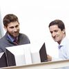 Two people in business attire talk in front of a desktop computer monitor.