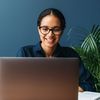 Woman in glasses uses a laptop on a desk with a notebook.