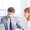 Two people sit in an office looking at a laptop.