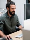 Person with beard in gray button-down shirt works on a desktop computer.