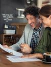 A smiling man and woman sit together at a wooden table in a workshop setting with chalkboard walls, woodworking equipment and a white sink, reviewing a report with charts and graphs. The man, wearing a light gray blazer over a checkered shirt, holds a pen and paper, while the woman, dressed in a green button-up shirt, leans in with a smile.