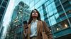 Young Indian woman in business casual attire, holding her phone and looking up at the sky outside while walking on street with glass buildings behind her
