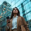 Young Indian woman in business casual attire, holding her phone and looking up at the sky outside while walking on street with glass buildings behind her