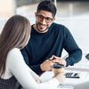 Two people converse while sitting at a small desk with a coffee cup, papers and a calculator on it.