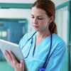 Healthcare professional wearing blue scrubs and a stethoscope uses tablet computer in a hospital hall.