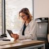 Doctor in white coat with stethoscope sits at sunlit desk reading a tablet.