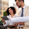 Two business colleagues outdoors smiling while reviewing a tablet.