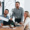 Four coworkers smile and discuss around a table with laptops and notebooks in an office meeting.