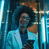 An African-American woman in spectacles smiles as she looks at her smartphone in a neon-light filled glass elevator.