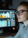 A young woman wearing spectacles is working in a dimly lit room. There is a monitor with bar charts displayed on it across from her. 