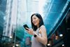 A low-angle shot of a young, smiling Asian woman looking at her smartphone as she holds a reusable coffee cup in an outdoor city setting.