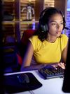 A young woman sits in a personal office or gaming room, wearing a gaming headset and typing on her keyboard. She is looking at her monitor screen.
