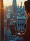 A woman stands by a window overlooking a city landscape as she is focused on her laptop.