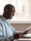 Prospective student smiling, sitting at desk and looking at a piece of paper as they prepare to enroll for school on a laptop.
