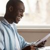 Prospective student smiling, sitting at desk and looking at a piece of paper as they prepare to enroll for school on a laptop.