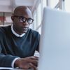 Person sits at laptop computer near a windowed wall.
