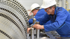 Two industrial workers in hard hats work on turbines.