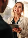 Medical professional wearing a white coat and stethoscope holds a tablet computer and smiles.