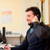 Person in business-casual attire in a small office speaking on a phone in front of a computer.