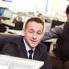 Two people collaborate on the content of a desktop monitor in an open office setting.