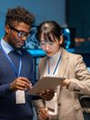 Two young professionals, an African-American man and an Asian woman, are looking at a tablet in a modern, dimly lit office. Both are wearing ID badges on lanyards.