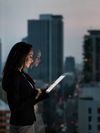 A young professional woman wearing a blazer stands beside a high-rise office window looking down at her tablet; outside the window you can see a city skyline with illuminated buildings.