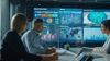 Business meeting with diverse team at a conference table, large screens displaying graphs, charts, and world map in a modern office setting.