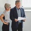 Two people stand in an office and look at paperwork.