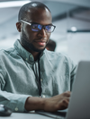 Person with glasses sits and types on a laptop.