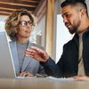 Two people stand, talk and look at information on a laptop.