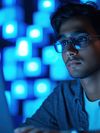 A young Indian man is working on a laptop against a blurred tech-themed background.