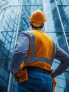 A man wearing a high-visibility vest, tool belt and a safety helmet over a shirt and jeans stands with his back facing the viewer, looking up at a completed high-rise office building. The reflective glass panels of the building create a dynamic pattern, emphasizing the scale and architectural design.