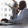 Two people in an office setting, one holding papers and another on a cell phone.