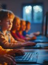 A group of young children are sitting at a row of desktop computers in a classroom setting, focusing on their screens.
