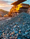 A close-up view of a pile of crushed rocks at a mining site, with a large dump truck, industrial machinery and the setting sun blurred in the background.