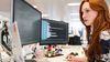 Person with red hair sits at a multi-monitor computer set-up in an office setting.