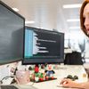 Person with red hair sits at a multi-monitor computer set-up in an office setting.