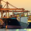 A large ship near port is pictured on the water and in front of shipping infrastructure.