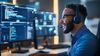 A smiling man wearing headphones and a headset happily works in front of two computer monitors.