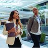 Two professionals in business attire with lanyards walking in a modern office atrium; one smiling.