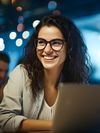 A woman in glasses smiles with her laptop in front of her against a blurred background of her colleagues working.