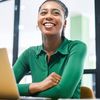 Person in a green blouse smiles while sitting in front of a laptop computer.