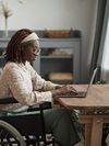 Person in wheelchair sits at a table in front of a laptop.