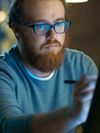 A bearded man wearing glasses intently draws on a display screen with a digital pen in a modern office.