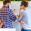 Two people in business casual clothing discuss and draw on a whiteboard.