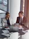 Two people sit and talk at an office table. One person shares information on a tablet.