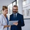 Doctor and suited professional discuss a tablet in a bright hospital hallway, wheelchair patient blurred in the background.