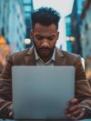 Focused young African American businessman using laptop on a busy city street at night.