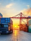 A large cargo truck carrying a blue shipping container is positioned in between tall stacks of red, blue and green containers in an industrial shipping port. In the background, red gantry cranes and a setting sun are visible.