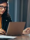A young woman wearing black-rimmed glasses, a yellow turtleneck and a leather jacket sits at a wooden desk, working at her laptop. Her phone, papers and a pen are spread out on the desk, and there is a gray curtain in the background.